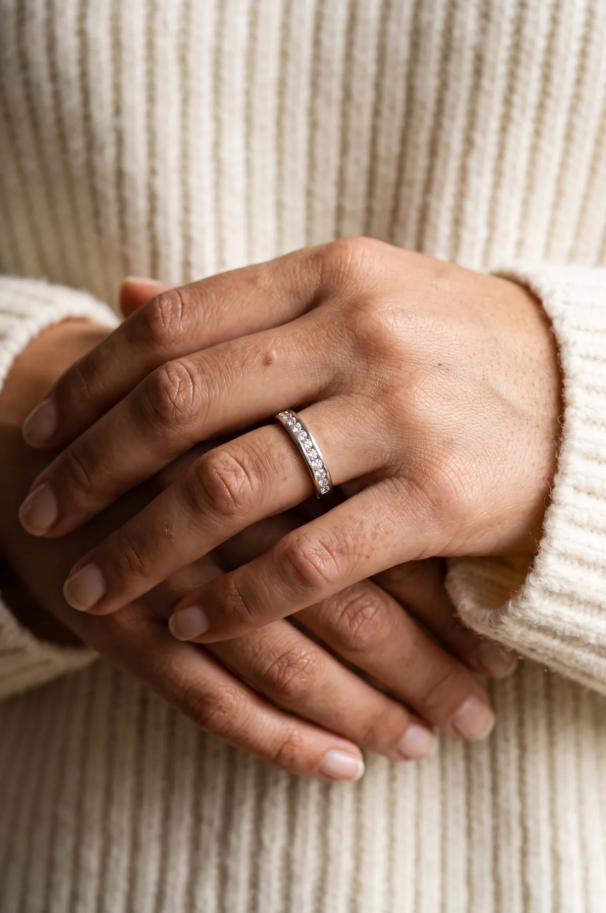 Two hands clasped together in the sleeves of a cream ribbed sweater, the upper hand wearing a band set with a row of channel-set diamonds