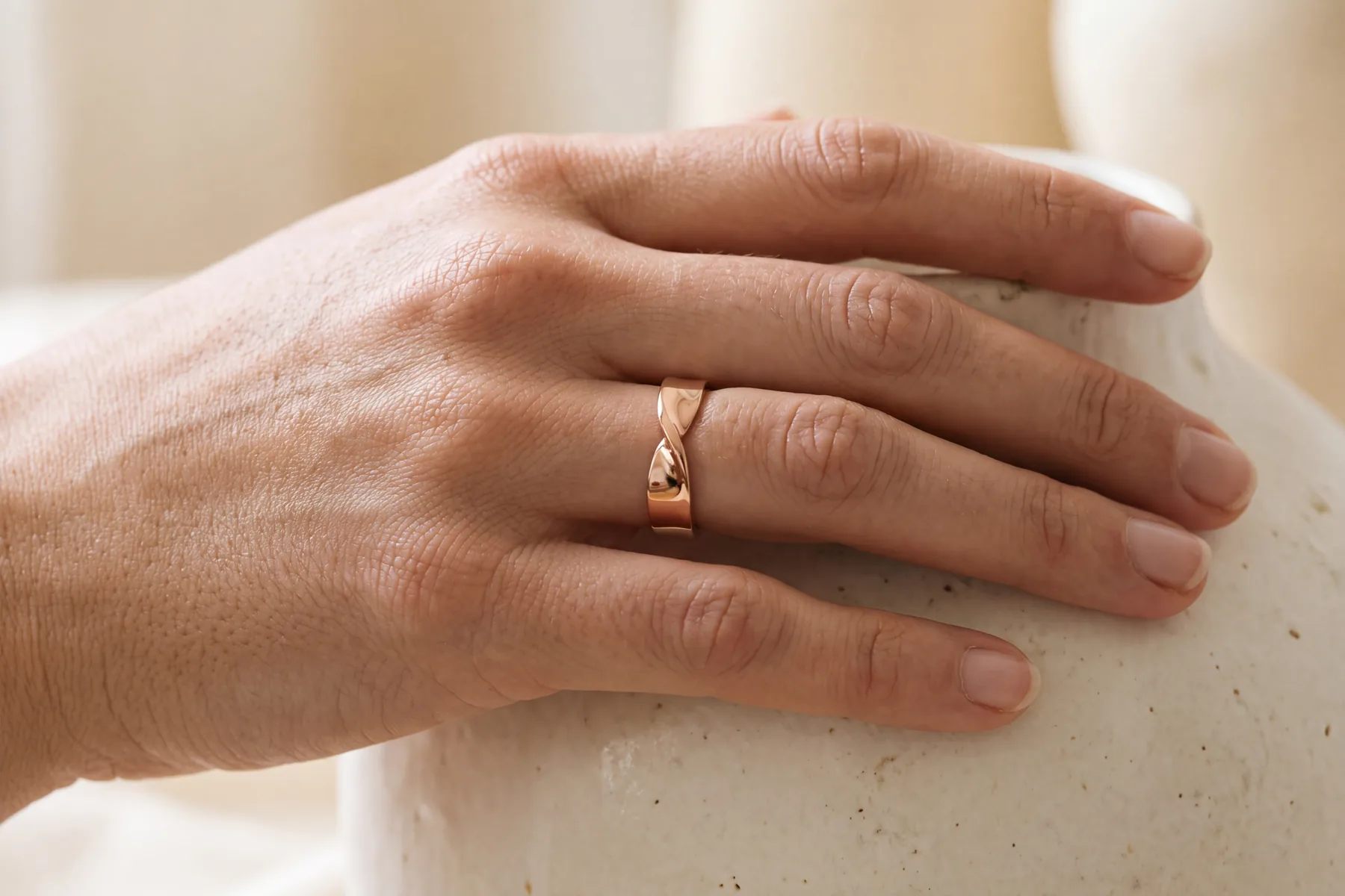 Close-up of a hand on a pale ceramic surface, wearing a slim rose gold band with a twisted rope profile